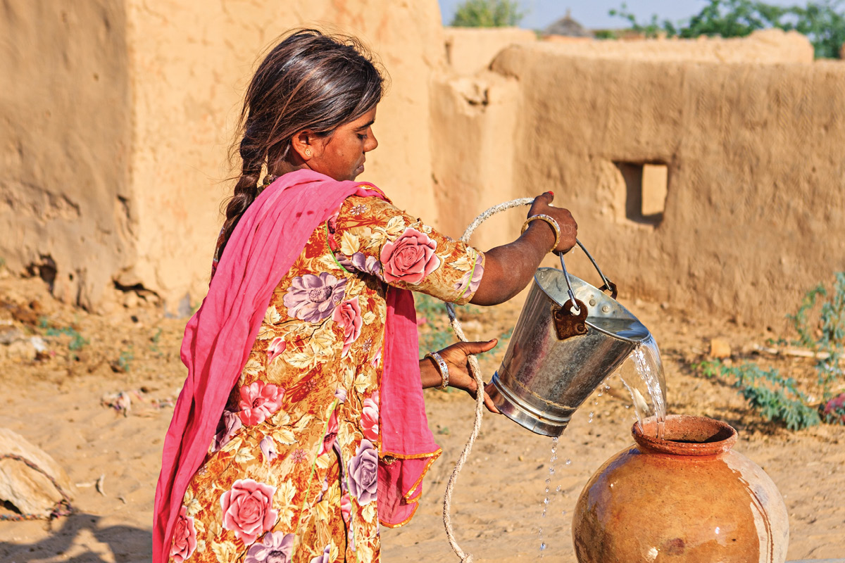 A brightly dressed girl pours water from a bucket into a clay pot