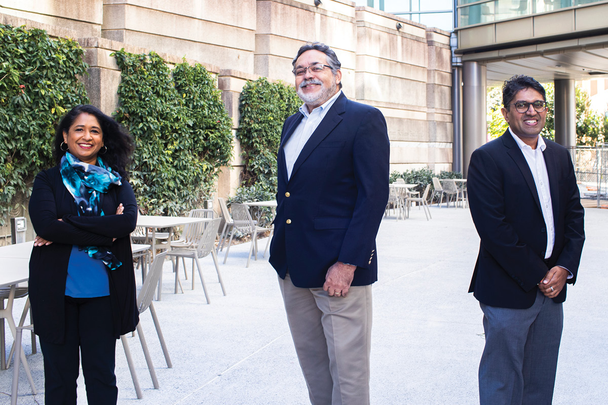 Jyothi Rengarajan, Kenneth Castro, and Neel Gandhi stand in a walled courtyard with tables in the background