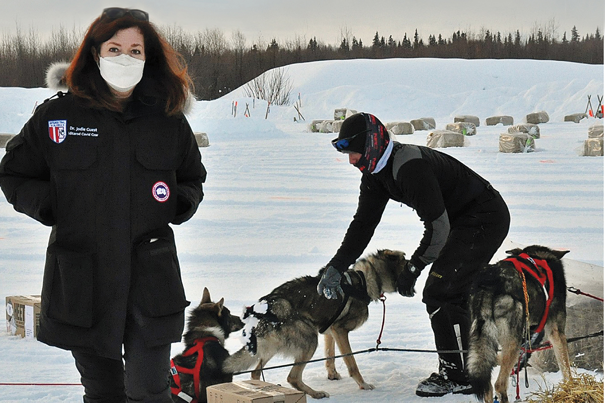 A masked woman wearing a blue parka stands in the snow. Behind her, a man wrangles sled dogs.