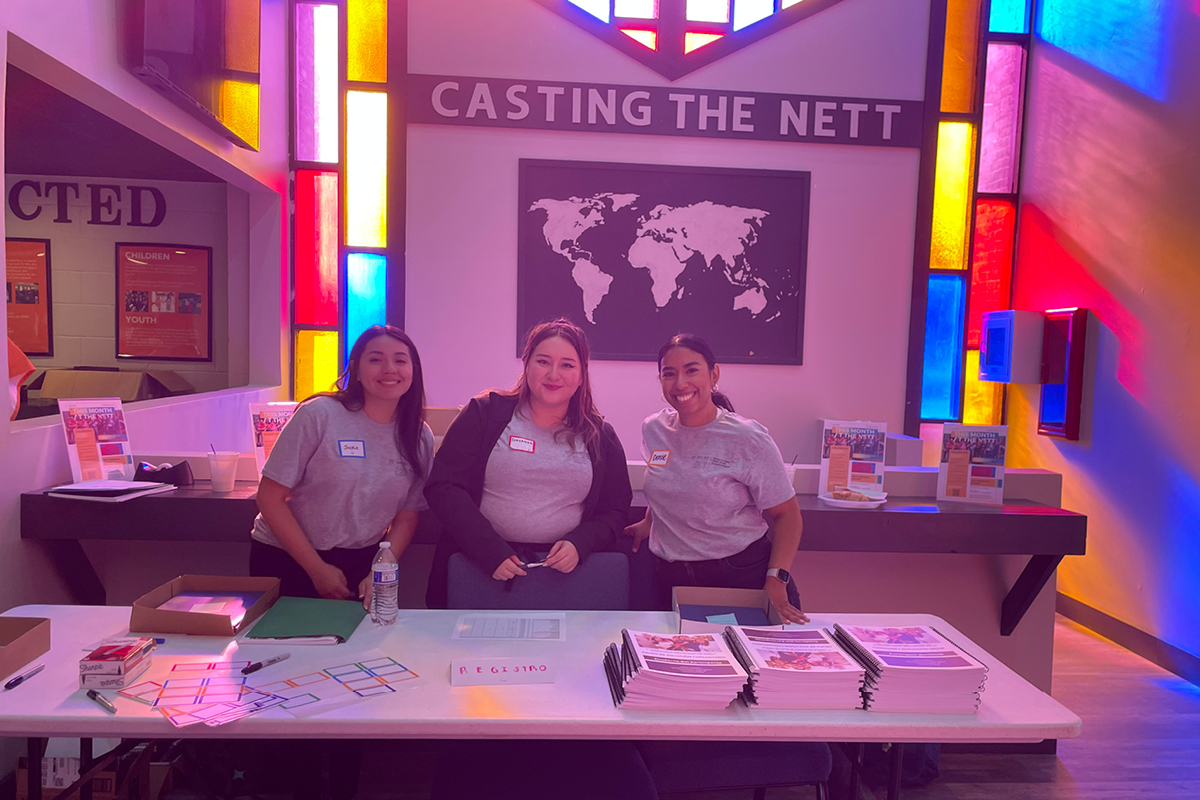 a photo of 3 women students smiling at a camera in a church in front of the table they set up for the event.